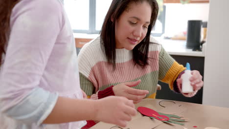 happy biracial mother and daughter sticking colourful cutouts in sunny living room