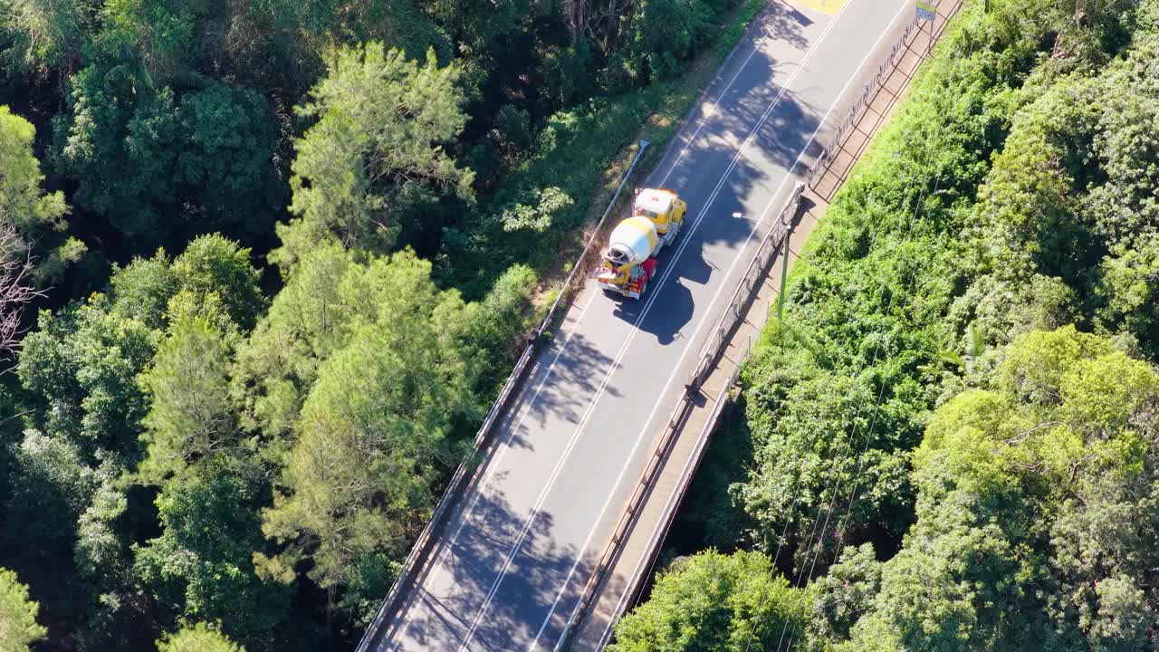 Aerial footage captures a cement truck driving through a lush forest road under bright sunlight