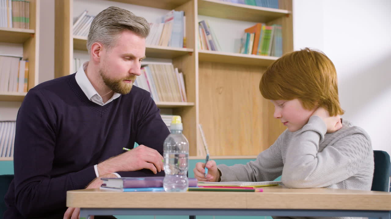 estudiante pelirrojo sentado en el escritorio en el aula de inglés mientras el profesor le responde algunas preguntas 1