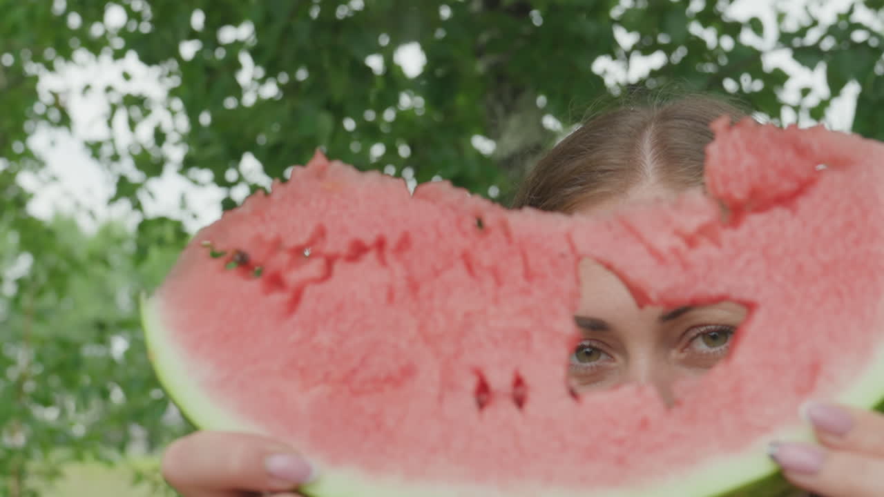 primer plano de una mujer caucásica mirando a través de una sandía, luz natural suave, fondo de jardín frondoso, manos sosteniendo una gran rodaja mordida con un agujero irregular, ojos visibles a través de la fruta, ambiente veraniego y divertido