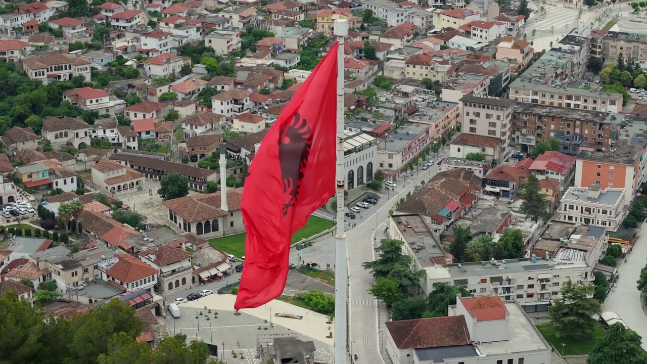 Sirkulært dronebilde rundt det albanske flagget over flislagte hustak, moskeer og minareter i Berat, Albania. Luftfoto av storslåtte osmanske bygninger.