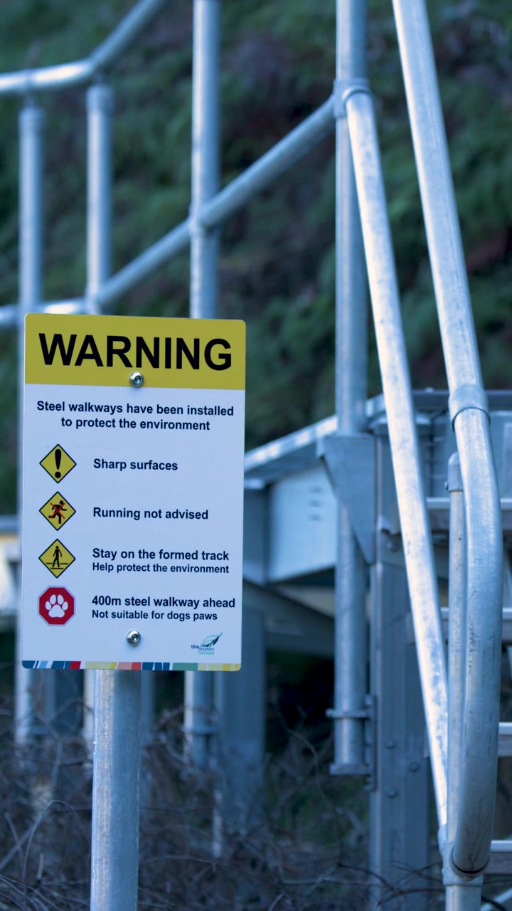 Camera pans down to reveal a yellow safety warning sign on metal hiking stairs.
