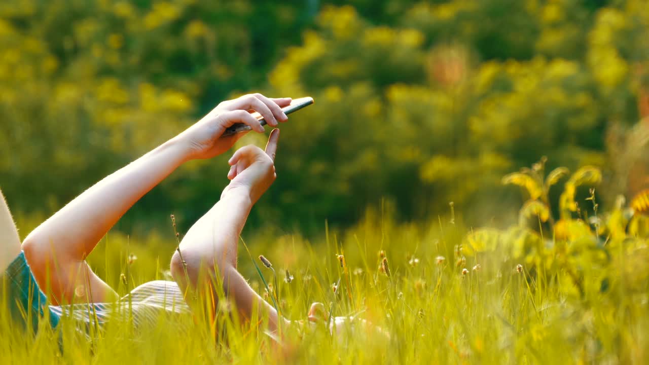 Happy Young Woman Lying on Green Lawn Uses Smartphone on Scenic Field at Sunset Background
