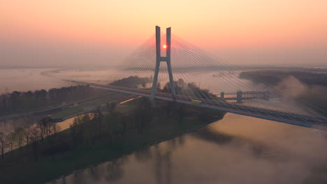 Aerial-shot-of-a-modern-cable-road-bridge,-over-a-wide-calm-river,-during-a-foggy-sunrise