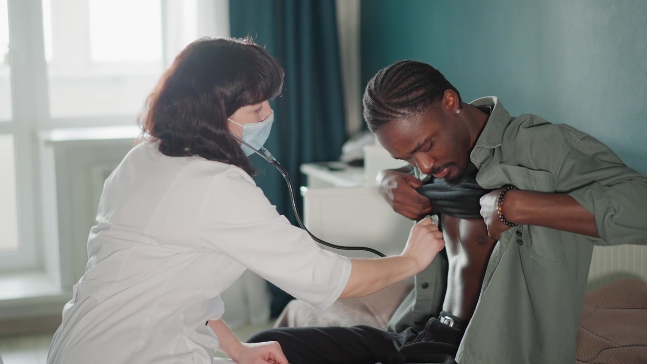 Black male patient home chest exam with white nurse using stethoscope and mask, calm livingroom setting, seated on chair, paramedic bag nearby, reassuring touch during vitals check, professional