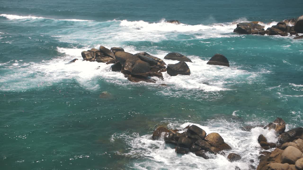 olas rompiendo contra una pila de rocas en el mar azul del parque tayrona, colombia