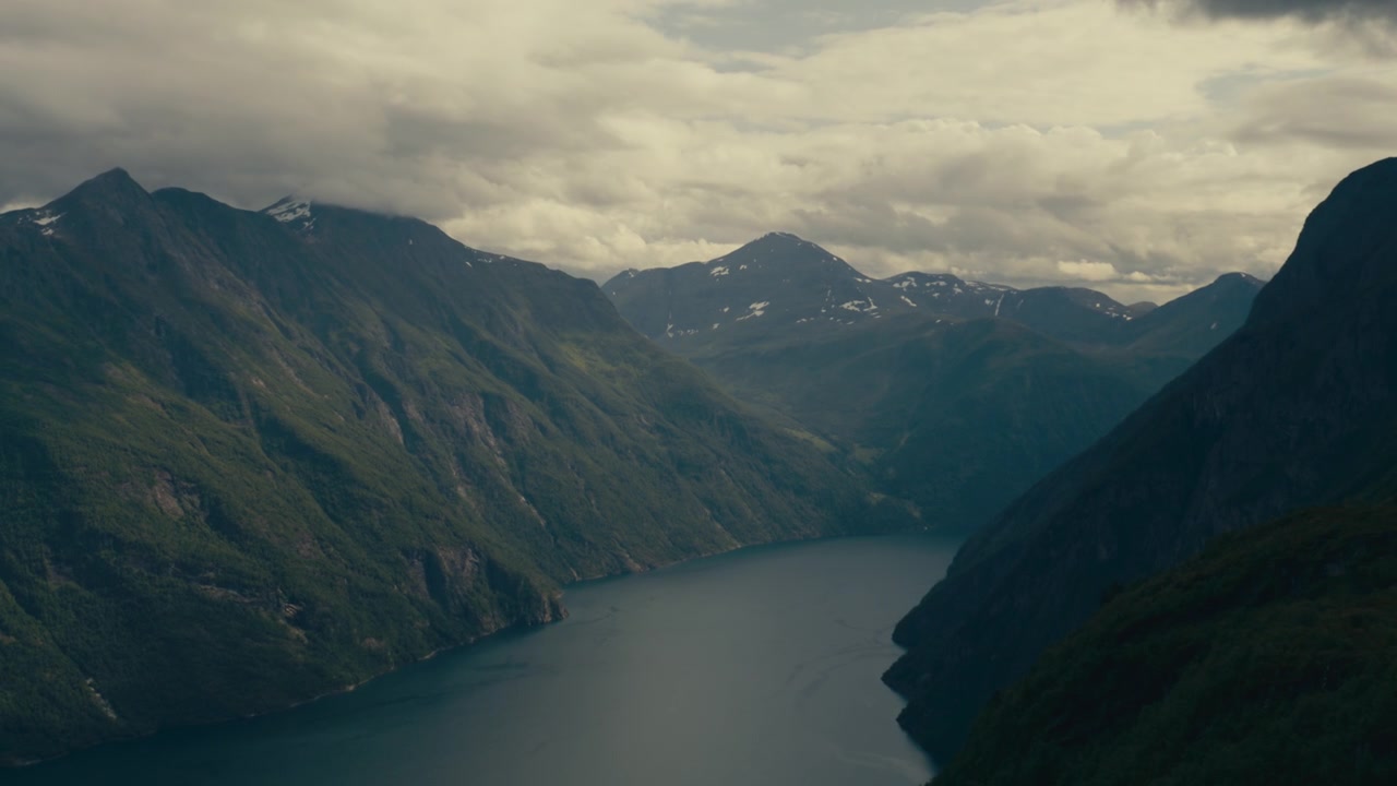 Langer norwegischer Naturfjord schlängelt sich zwischen dunklen Berghängen unter einem bewölkten Himmel, von einem hohen Aussichtspunkt am Hang aus gesehen