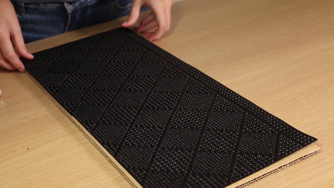 Hands pressing a black rubber mat onto a cardboard surface, testing adhesion. Bright lighting, close-up view, steady camera