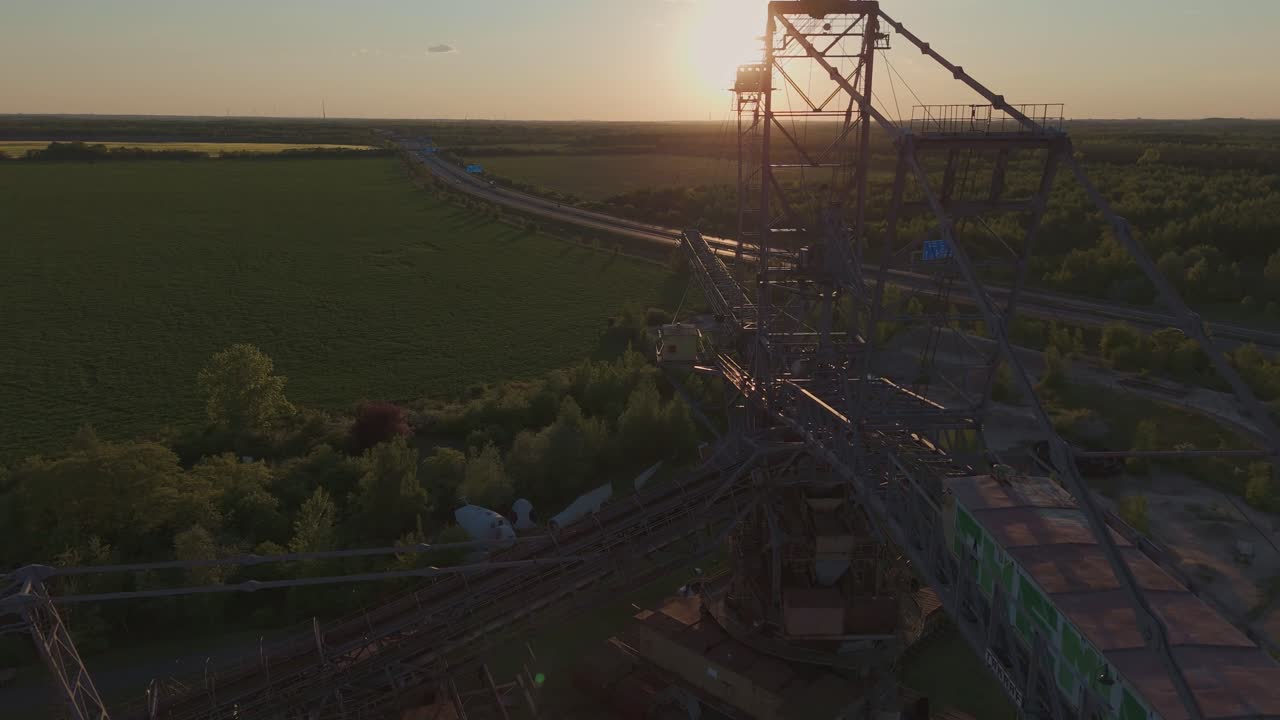 Drone footage of a massive stacker machine near Leipzig, captured at sunset. The industrial structure rises above green fields with a distant highway, blending technology and nature in golden light.