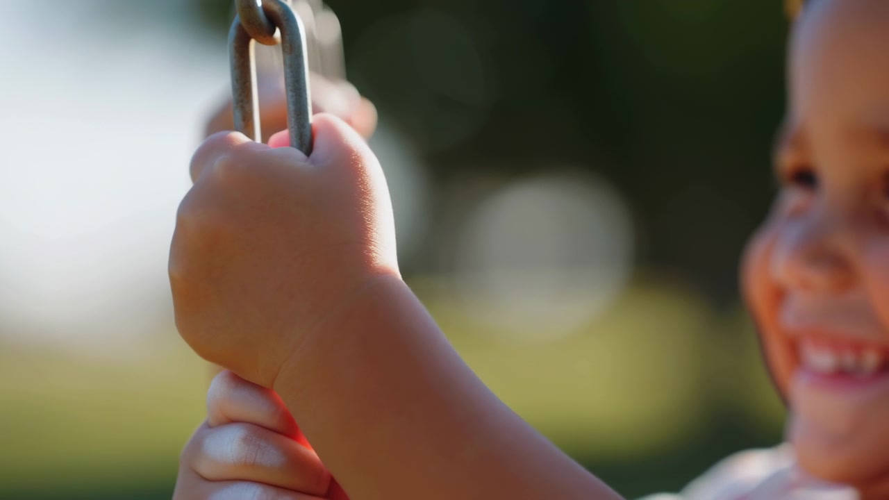 Child hands grip weathered metal swing chain in close-up, with slight upward repositioning of fingers around links, sunlit playground setting, soft background blur, metal texture and partial face at