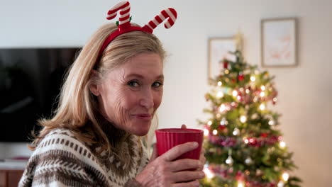 smiling woman wearing festive headband enjoying hot drink by christmas tree