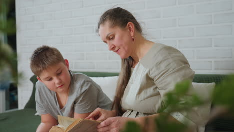 mom flipping book page while son in grey shirt closely observing on green couch with white pillows, indoor cozy room with soft natural light, bonding moment over old vintage book