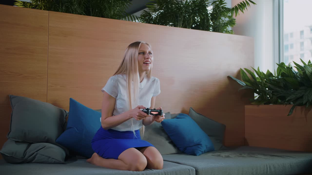 Woman gaming and relaxing in modern office. Young formal woman in skirt chilling on sofa in contemporary office recreation area and playing videogame with gamepad
