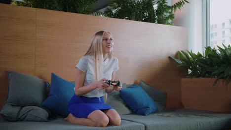 woman gaming and relaxing in modern office. young formal woman in skirt chilling on sofa in contemporary office recreation area and playing videogame with gamepad