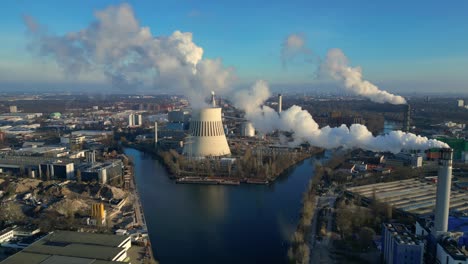 Aerial view of a thermal power plant emitting smoke on a sunny day, with a river and a city in the background. Dramatic aerial view flight static tripod hovering drone