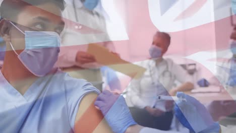 Animation-of-flag-of-uk-over-female-doctor-in-face-mask-giving-covid-vaccination-to-male-patient