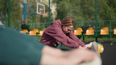 close-up of lady in green hoodie working out outdoors with two other ladies stretching in background, featuring basketball court and fitness training in sports arena