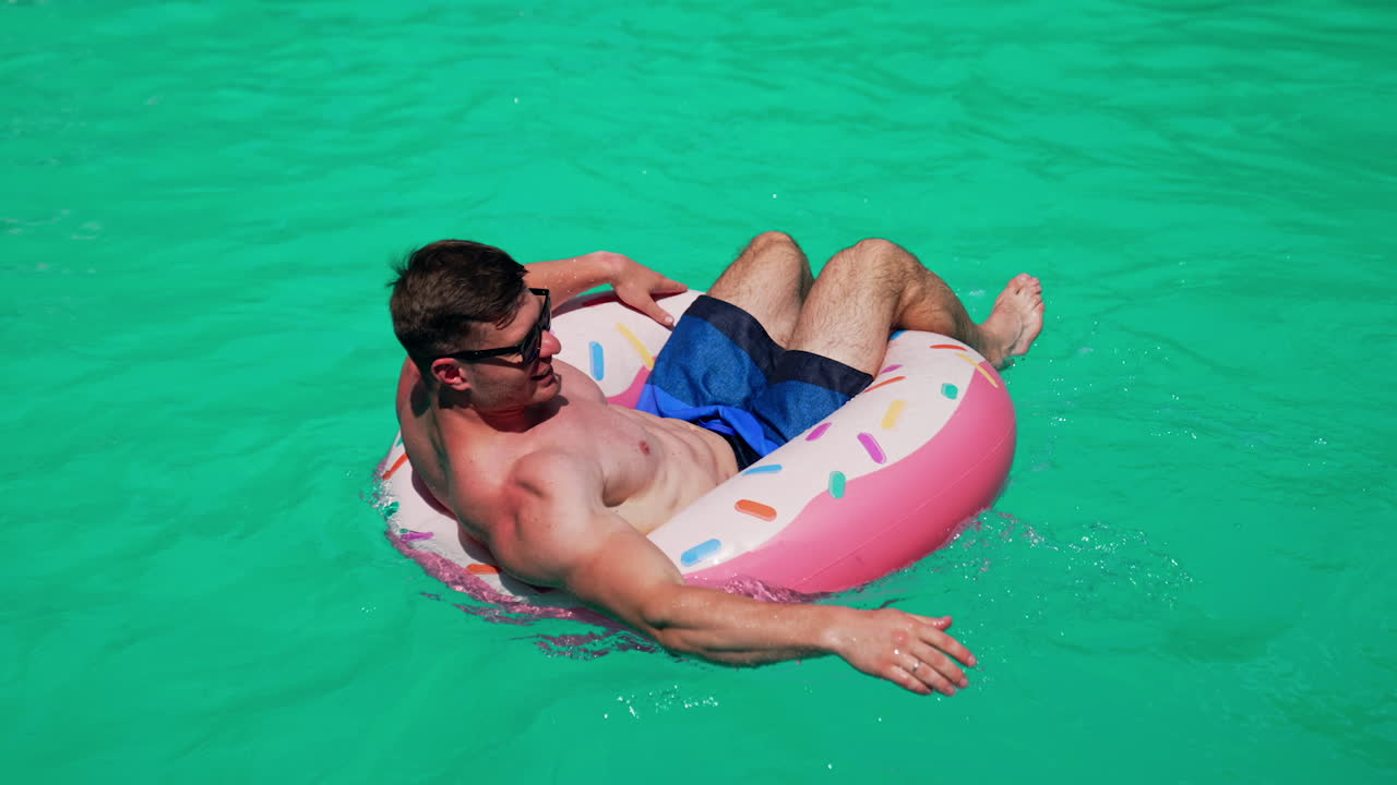 Man wearing sunglasses resting in the sun. Man sits in the inflatable circle in the pools rowing with hands.
