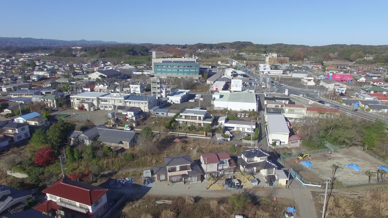 この動画は、日本の郊外の町を広大な空撮視点で捉えています。伝統的および現代的な住宅建築物が、様々な商業施設や道路と混在している様子を紹介しています