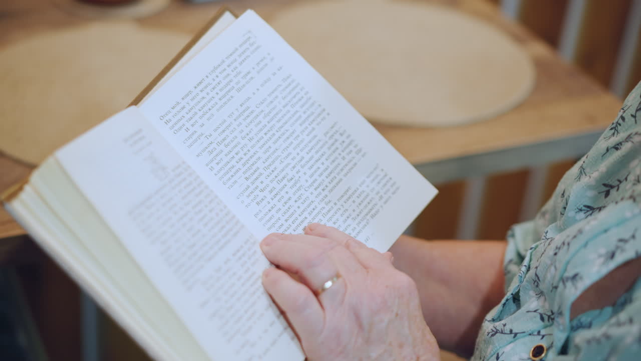Elderly lady studies open pages quietly. White elder woman siting at table immersed in reading activity. An aged white grandmother peacefully reads book at her dining table during daylight hour
