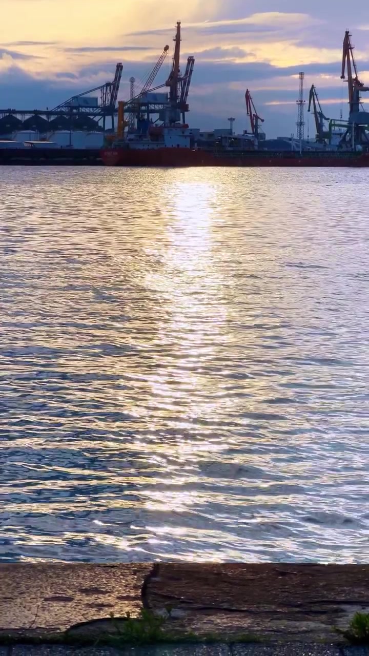 Solnedgang over havnen med kraner. Udsigten over havnen viser vand, der reflekterer solnedgangen. Kraner er synlige i baggrunden mod himlen.