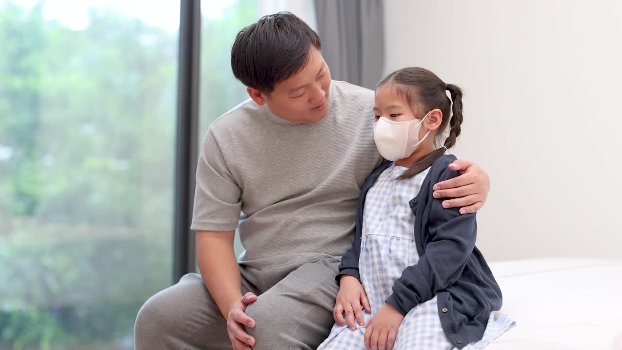 An Asian father gently comforts his masked daughter in a bright, modern room. Soft natural lighting, steady camera, and a supportive, empathetic mood