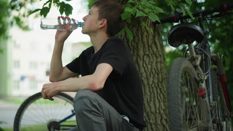 young boy drinks water while resting under a tree after a bicycle ride, the bicycle is parked close by, surrounded by lush greenery. blur view of vehicles passing in the background