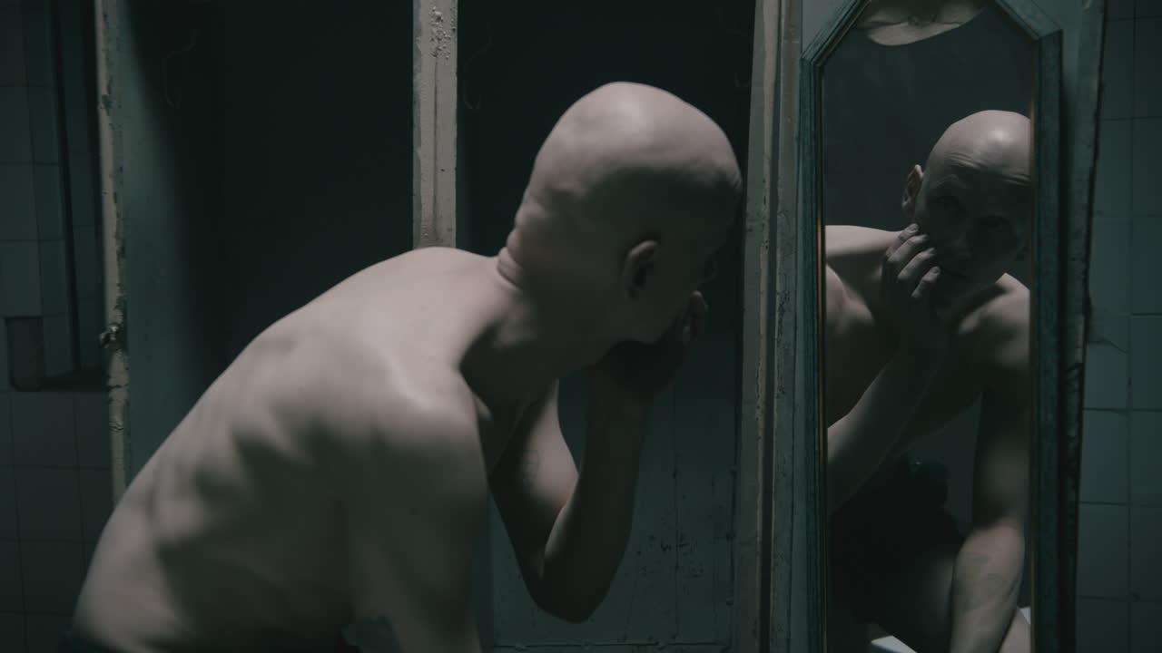 Pale bald skinny man looks into the mirror in the creepy old bathroom at night