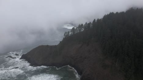 A-fog-shrouded-tree-lined-jagged-ridge-juts-out-into-a-harsh-rugged-coastline,-aerial