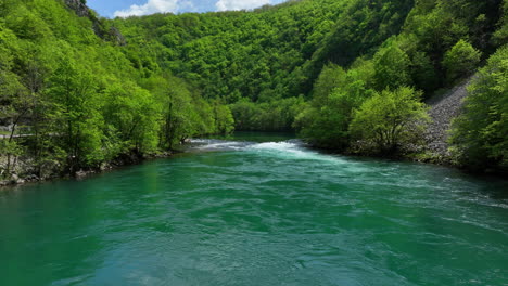 Low-aerial-shot-of-a-clear-green-river-surrounded-by-lush,-newly-leafed-trees-in-early-spring,-under-a-sky-with-scattered-clouds