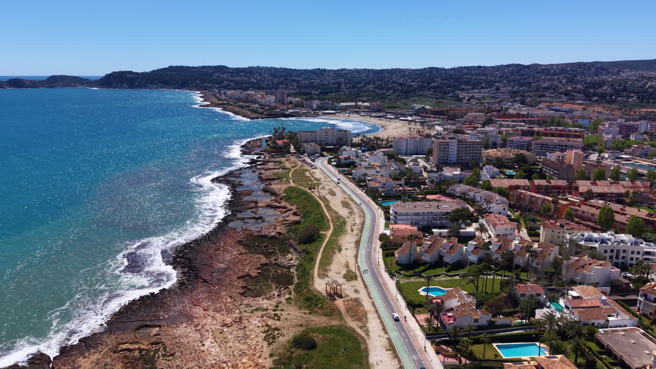 Città costiera di Denia a Valencia, Spagna, che mostra il Mar Mediterraneo turchese, la costa rocciosa, la strada costiera, gli edifici residenziali e la spiaggia sabbiosa che si estende in lontananza.