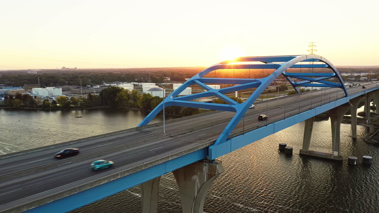 The Leo Frigo Memorial Bridge glows in the warm hues of sunset as cars cross its blue steel frame over the Fox River, reflecting golden light across the calm rippling water
