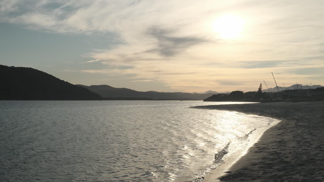 Static 4K shot at golden hour on a calm beach. Gentle waves roll onto the sand. Layers of distant mountains frame the scene, with weathered old boats washed up along the shoreline