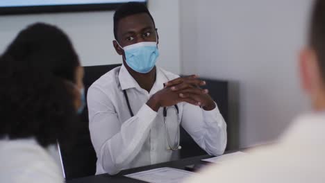 african american male doctor sitting at table in meeting room wearing mask speaking