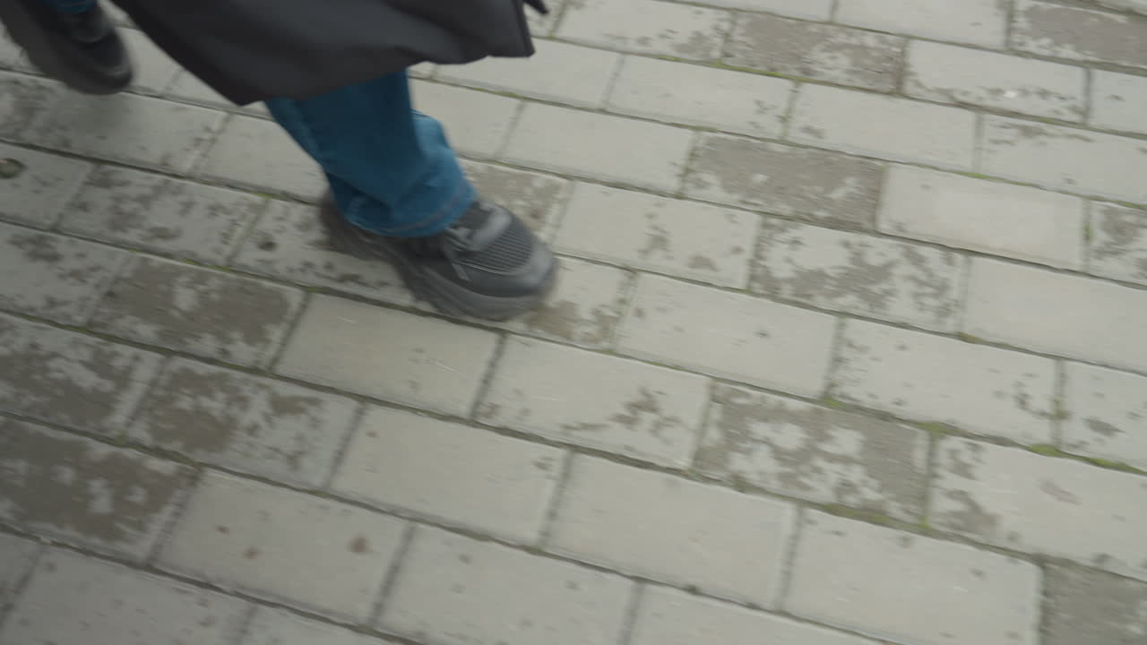 wet pavement footsteps shoes walking through light rain on brick sidewalk lowerbody frame shows blue jeans and dark coat, rubbersoled shoes stepping through shallow puddles beside metal railing