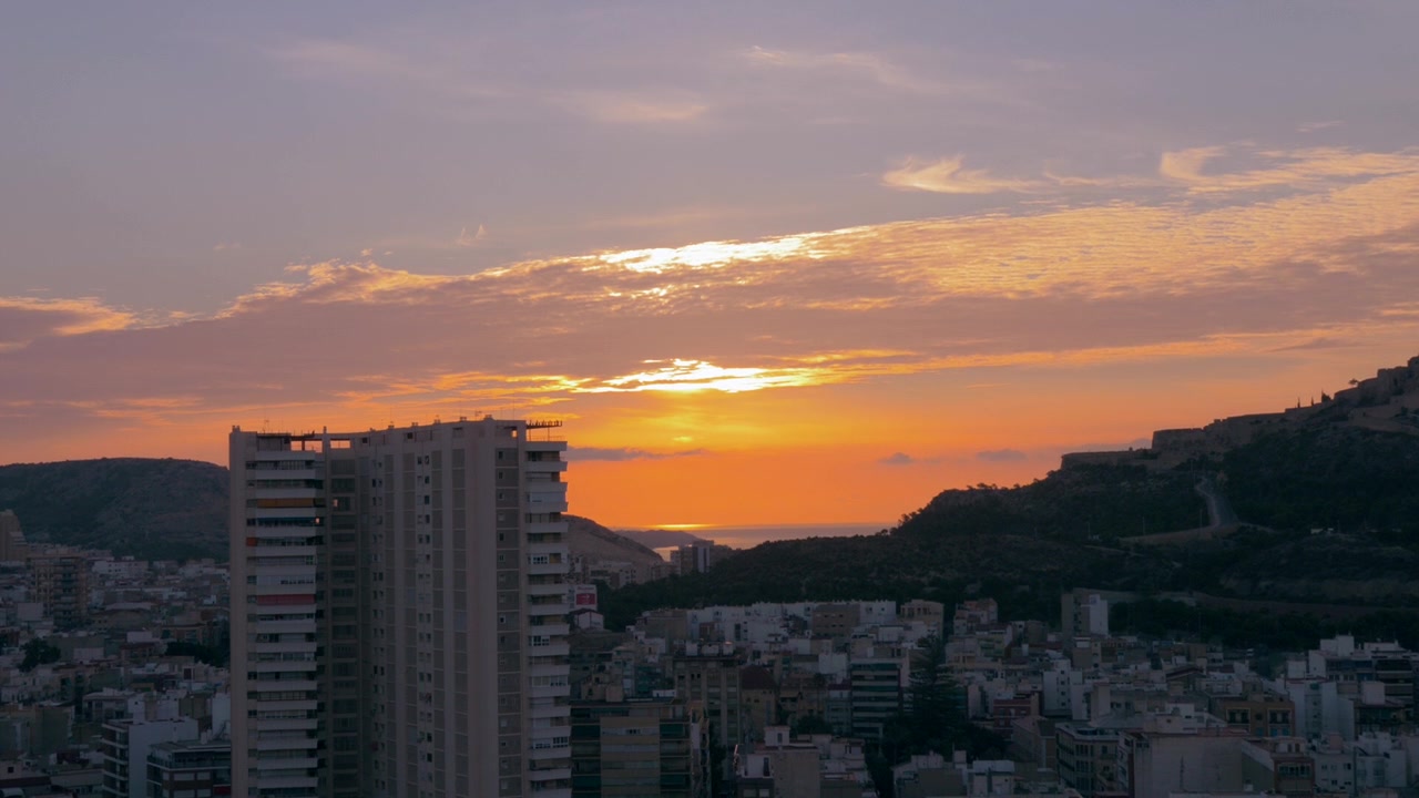 Bela paisagem urbana montanhosa, ao fundo vemos o mar. Céu laranja e nublado. Viagem na Europa, cidades com mar, Espanha, Alicante