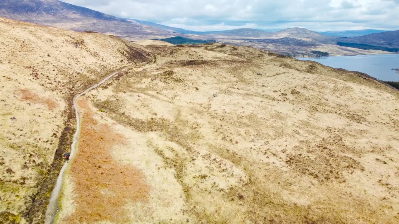 Aerial drone footage flying forward above a person hiking on a rural desolate and small road in scotland grassy yellow and brown hills and mountains. Large blue lake and mountains visible in the back.