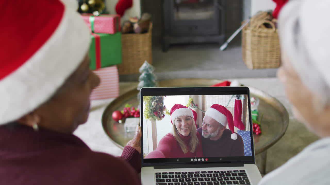 amigas mayores diversas usando una computadora portátil para una videollamada de navidad con una familia sonriente en la pantalla