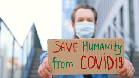 caucasian man wearing facial mask while holding save the humanity from covid" signboard and looking at camera in the street"