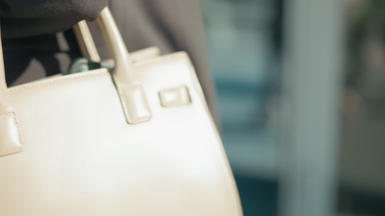 Close up partial view of woman in business outfit holding handbag walking by with her reflection visible on glass background, sunlight casting soft shadow on bag