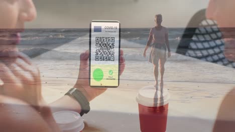 Woman-holding-a-smartphone-with-qr-code-on-screen-against-caucasian-woman-walking-on-the-beach