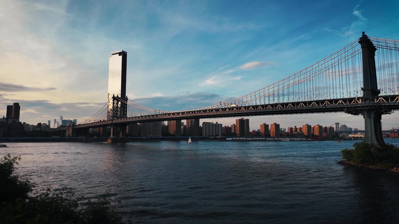 Ampia veduta statica del Ponte di Manhattan che attraversa l'East River con barche di passaggio, calda luce del tramonto e lo skyline di New York sotto nuvole serali colorate.