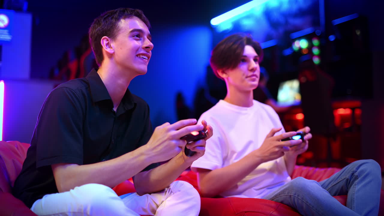 Two teen friends are playing a game console using gamepads, talking, laughing and pushing each other while sitting on bean bags. Blue and red illumination