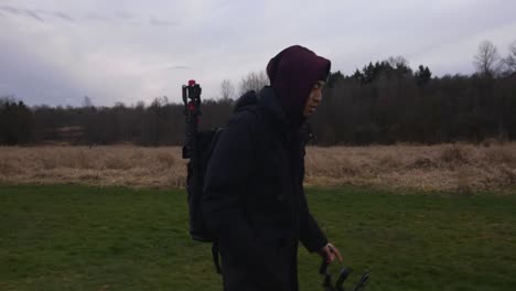 side view of a man walking on a grass field in a park with a back pack filled with equipment tripod with a hood and toque on