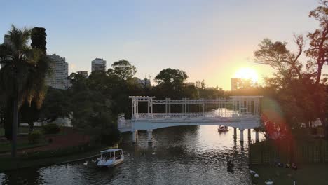 dolly aéreo de barcos navegando bajo el puente blanco en los jardines rosedal en el barrio de palermo al atardecer, buenos aires
