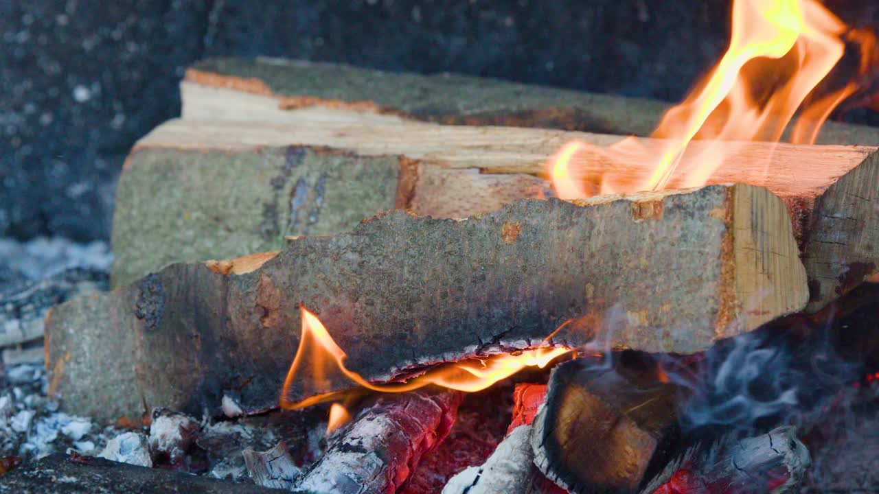 Stacked firewood burns with visible flames, glowing embers, and smoke in a steady close-up shot