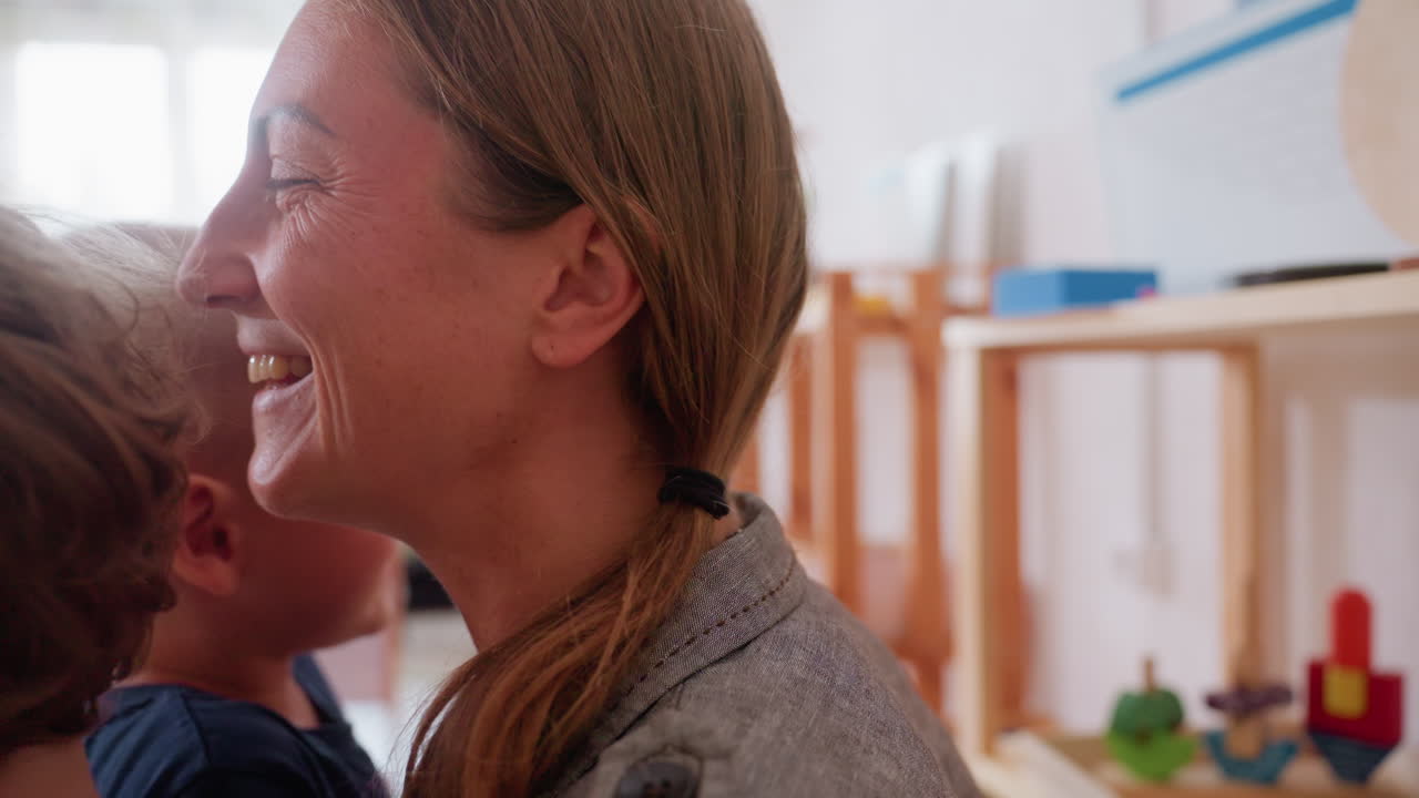 Smiling educator embraces two small children warmly in bright classroom surrounded by wooden shelves and toys, expressing love, care, and emotional connection while creating safe supportive