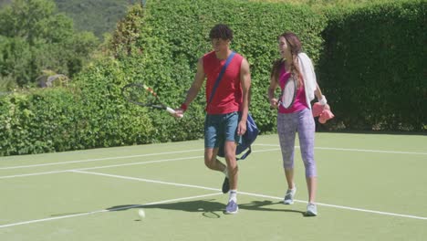 feliz pareja biracial caminando con bolsas y raquetas de tenis en el jardín en un día soleado