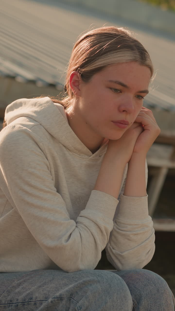 Close-up of thoughtful young woman in casual hoodie and jeans sitting on rustic stadium bleachers, resting hands on chin with a pensive and reflective expression