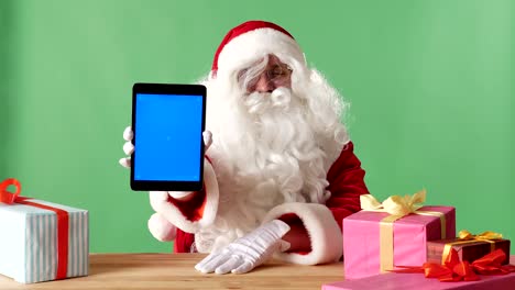 smiling santa claus showing tablet with blue screen in camera, gifts on the table, chromakey in the background